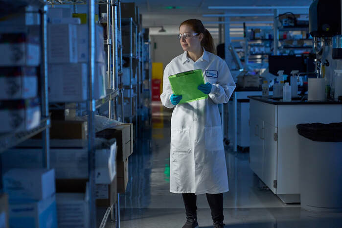 Scientist wearing white lab coat with a Biogen name tag, safety glasses, and gloves holding a green clipboard