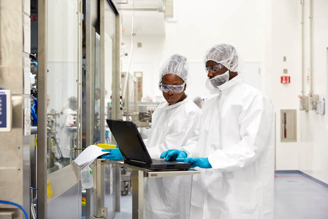 Two scientists wearing full-body suits, hair nets, gloves, and goggles work together at a laptop on a mobile workstation in a sterile lab